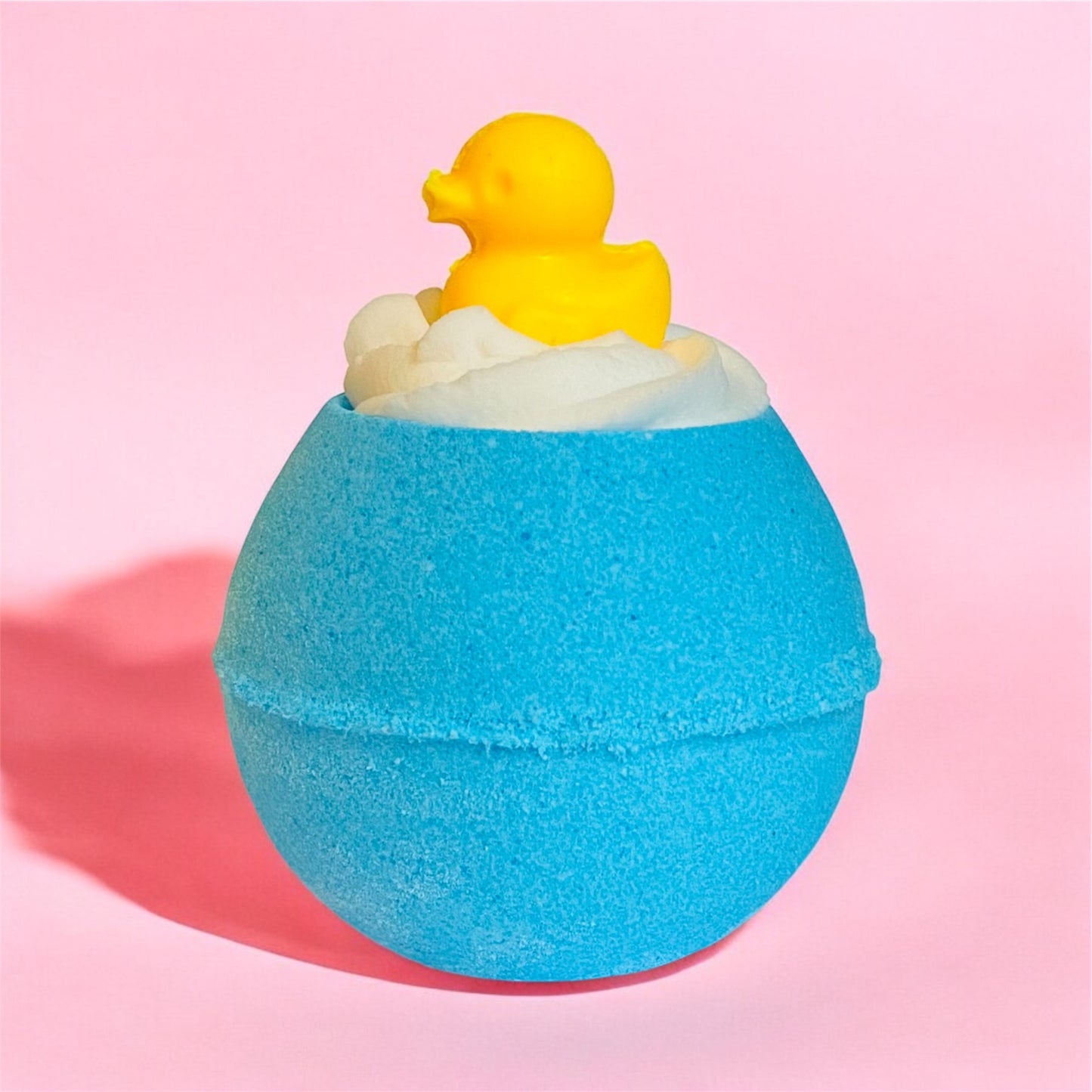 A  blue bath bomb  with a white whipped bubble bath frosting on top and a yellow duck shaped soap on top, on a pink background. 
