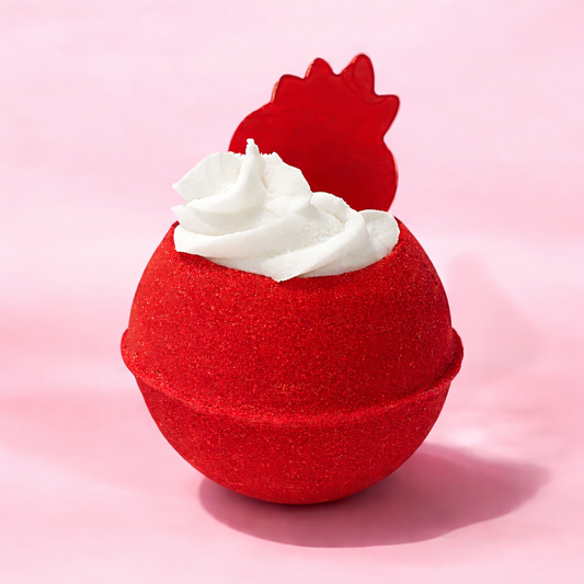 Red bath bomb with white whipped bubble bath frosting on top and a strawberry shaped soap on top, on a pastel pink background.