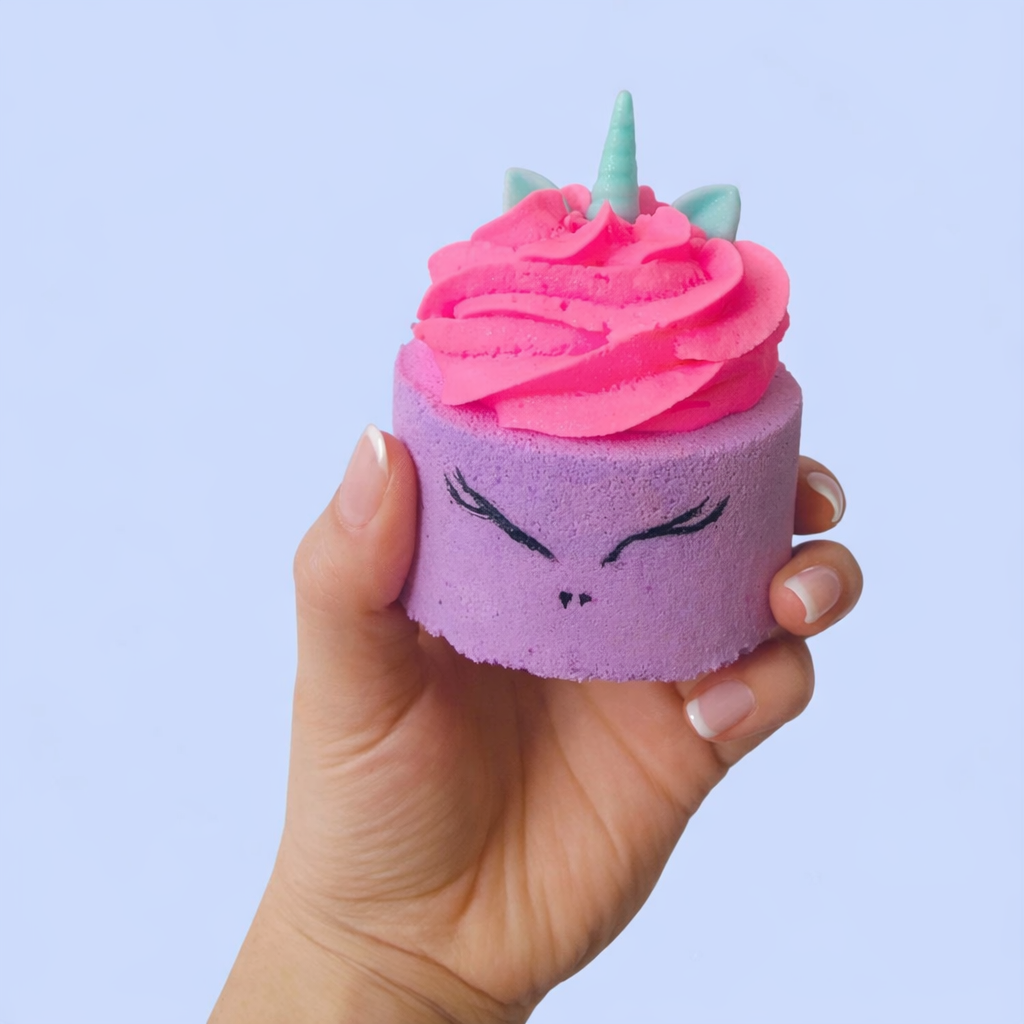 a purple cylinder shaped bath bomb with a pink whipped bubble bath frosting and a mini unicorn horn and ears shaped soap on top, on a pastel purple background.