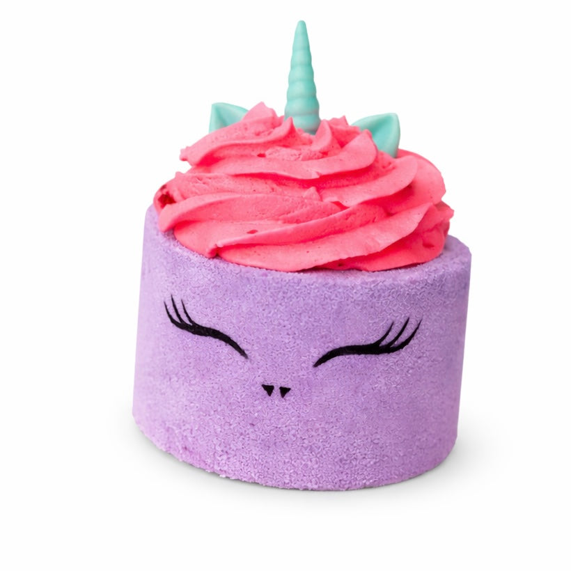 A purple cylinder shaped bath bomb with pink whipped bubble bath frosting on top and mini unicorn ears and horn shaped soap on top on a white background.