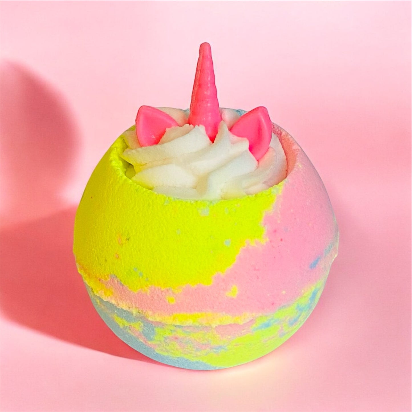 A multicoloured bath bomb with a white whipped bubble bath frosting on top and a unicorn horn and ears shaped soap on top, on a pastel pink background.