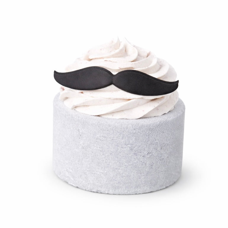 A grey cylinder shaped bath bomb with white whipped bubble bath frosting on top with a mini moustache shaped soap on top on a white background.