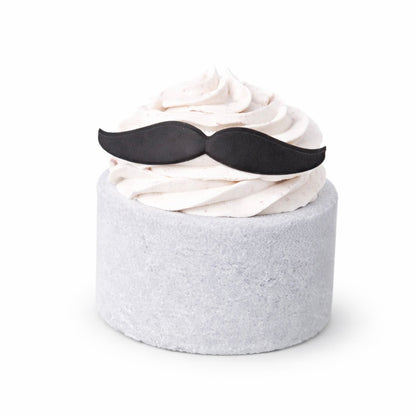 A grey cylinder shaped bath bomb with white whipped bubble bath frosting on top with a mini moustache shaped soap on top on a white background.